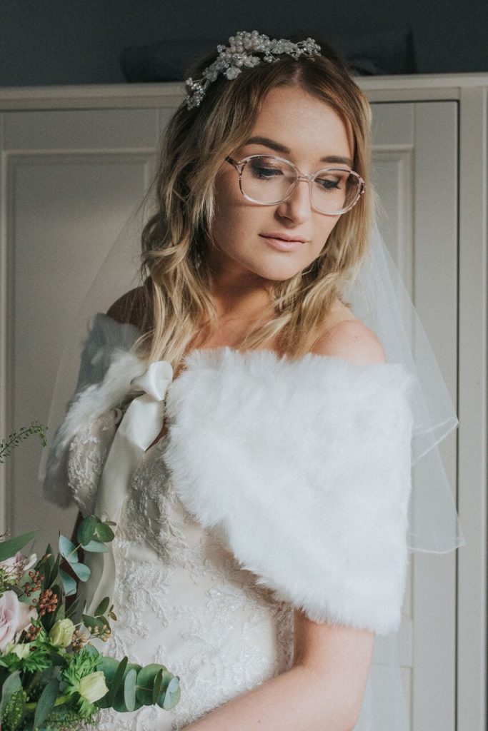 close up of bride wearing an ivory furry shrug, glasses, and delicate pearl headpiece. 