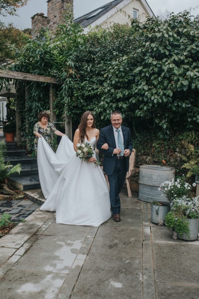 Bride arriving to wedding ceremony on the terrace accompanied by her father with her mother holding her long train. Her dress is a plain ivory strapless a-line gown and she hold her bouquet in her free arm