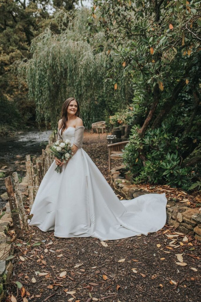 Bride on the riverwalk beneath the willow tree. Her dress is a plain silk-satin a-line gown and she is holding her ivory rose bouquet