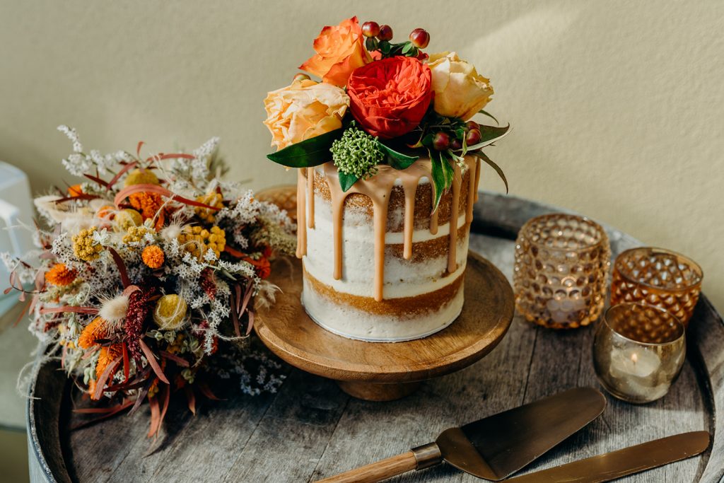 small semi naked wedding cake on a wooden cake stand decorated with drop and fresh roses on a wooden barrel with dried flower bouquet and candles