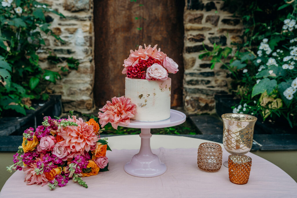 mini wedding cake with gold leaf on pink milk glass cake stand decorated with fresh pink flowers in front of a stone wall rustic backdrop
