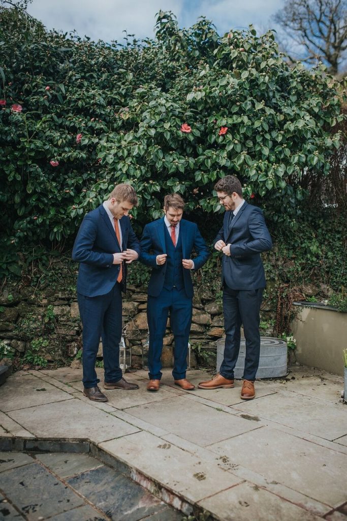 groom and two friends in navy suits with orange ties doing up their jacket buttons