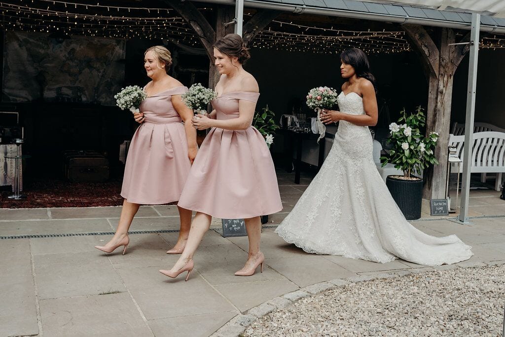 bride walking to ceremony with two bridesmaids in pink knee-length dresses walking in front of her,