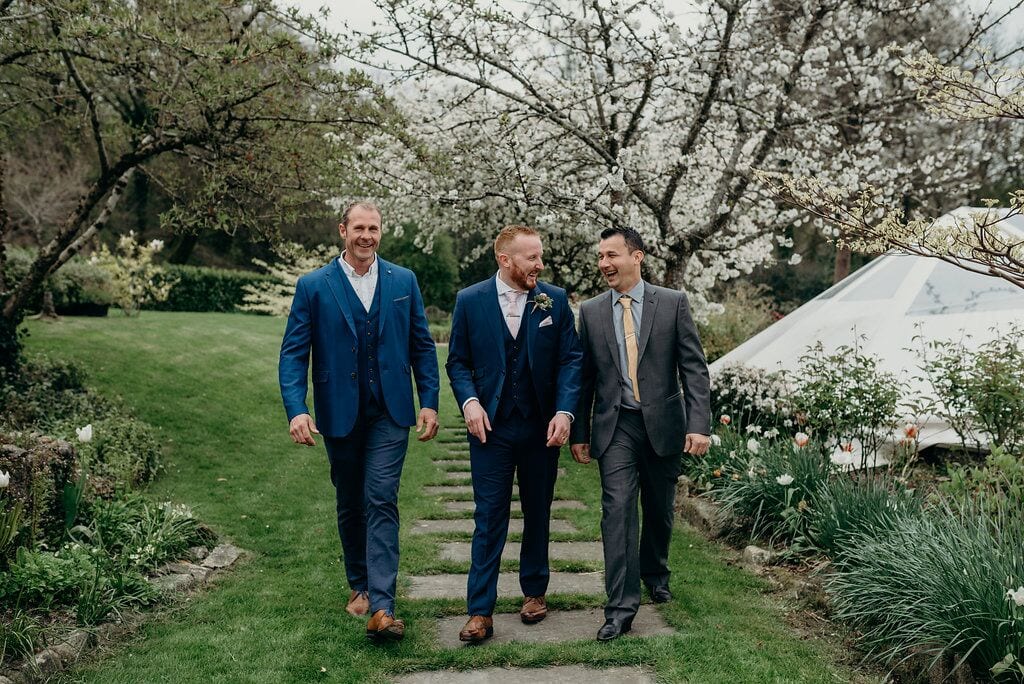 groom and two friends walking to ceremony beneath the blossom tree by the ponds.