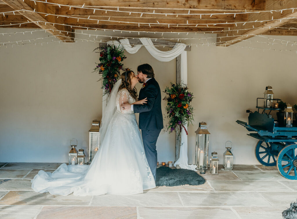 bride and groom stood on a black sheepskin rug in winter sun in front of a draped arch with dark flower sprays and steel lanterns