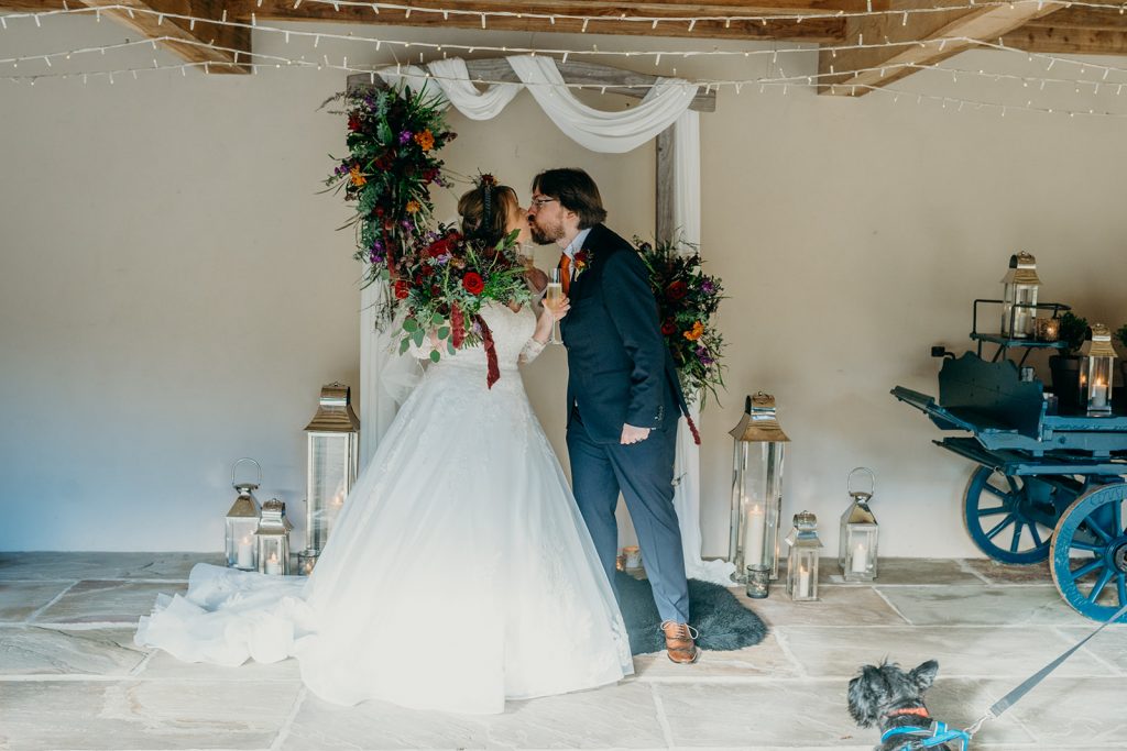 bride & groom kissing under a wooden arch with draped fabric and flowers and lanterns