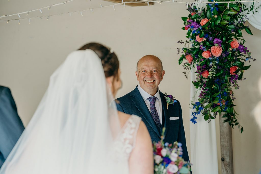 groom smiling as bride comes into wedding ceremony with flower spray backdrop