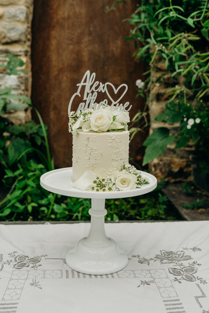 white iced mini wedding cake on a white milk glass cake stand decorated with fres white flowers in front of a corten steel water feature 