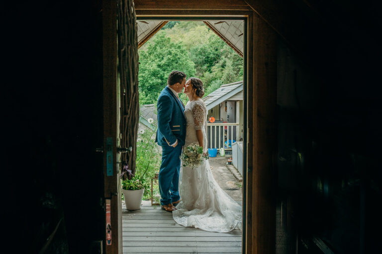 view through a wooden door of full view of bride in white lace gown with white bouquet and groom wearing navy suit facing each each