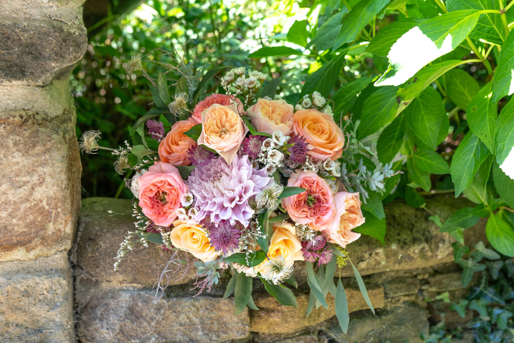 outdoor shot of a bridal bouquet with yellow and pink roses and purple dahlias set on a stone wall with green foliage in background