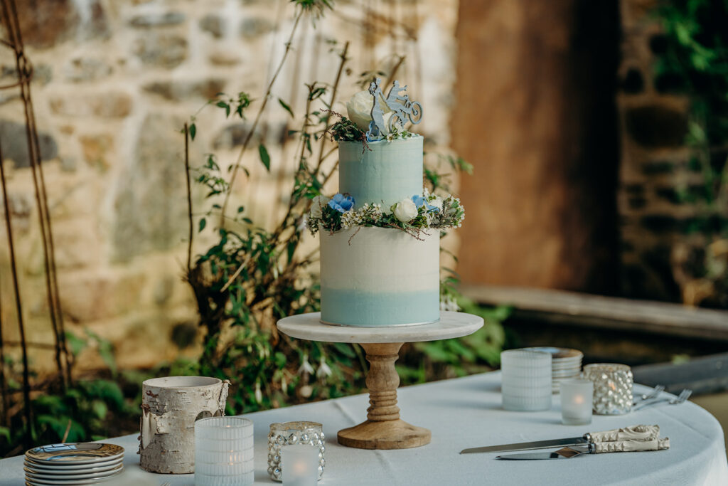 blue ombre elopement wedding cake on wooden and marble cake stand decorated with cake topper and fresh blue and white flowers with white and silver tealights in front of a stone wall