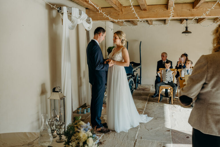 bride and groom getting married in front of a wooden fabric draped arch in rustic barn with wedding guests seated on wooden chairs
