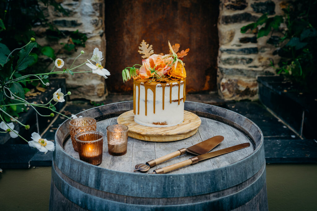 mini wedding cake decorated with caramel drip and autumn coloured flowers and leaves on a round wooden board set on an oak barrel in front of a rustic wall