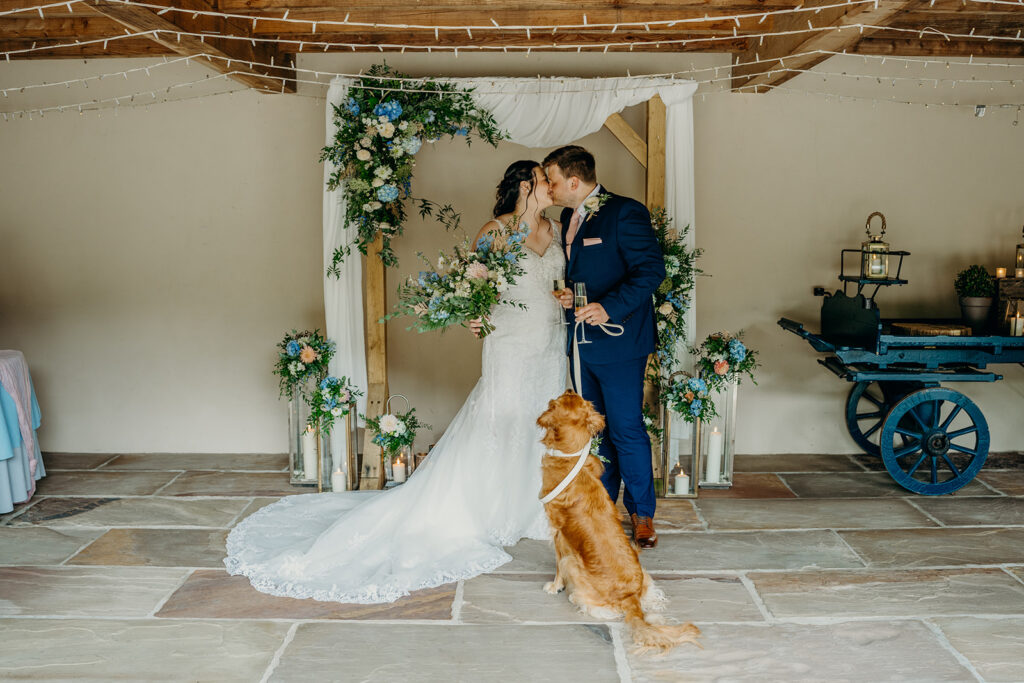 newly wed couple kissing in front of wedding ceremony arch decorated with pink and blue flowers with gold retriever dog gazing at them