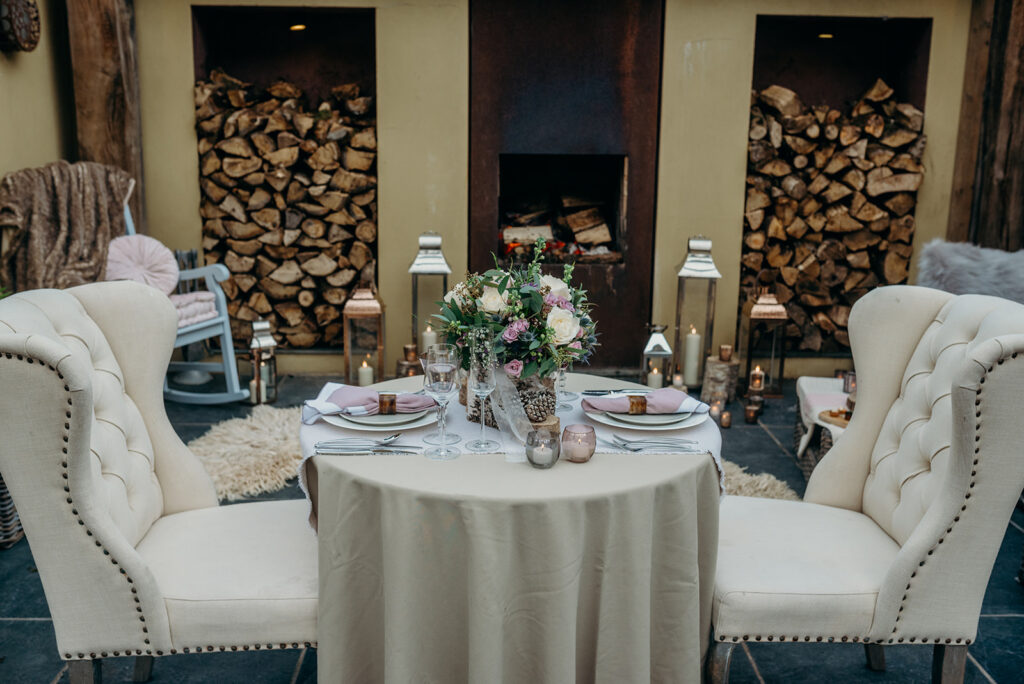 wedding table in cream and pink set with two cream upholstered chairs in front of a fire with log stacks and lanterns 