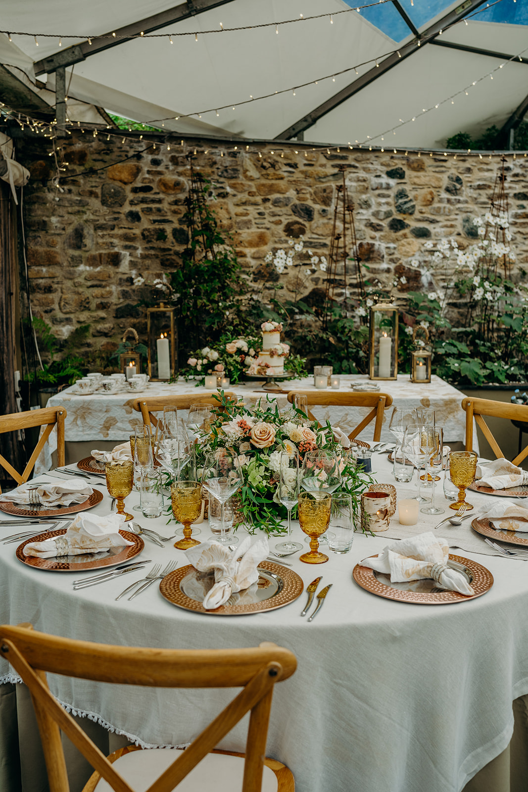 round wedding breakfast table with cake table in the background dressed in rustic table linen and rose gold chargers and amber water goblets 