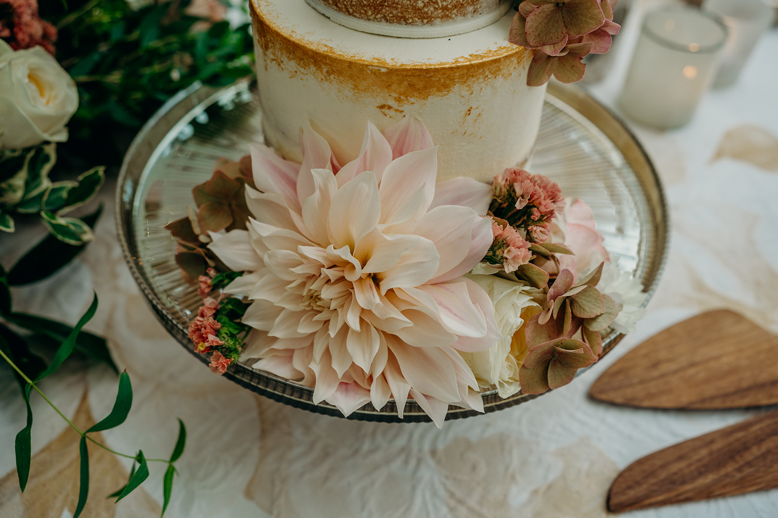close up of cafe au lait dahlia decorating a two tier wedding cake on a dark lustre glass cake stand with wooden cake knife and slice