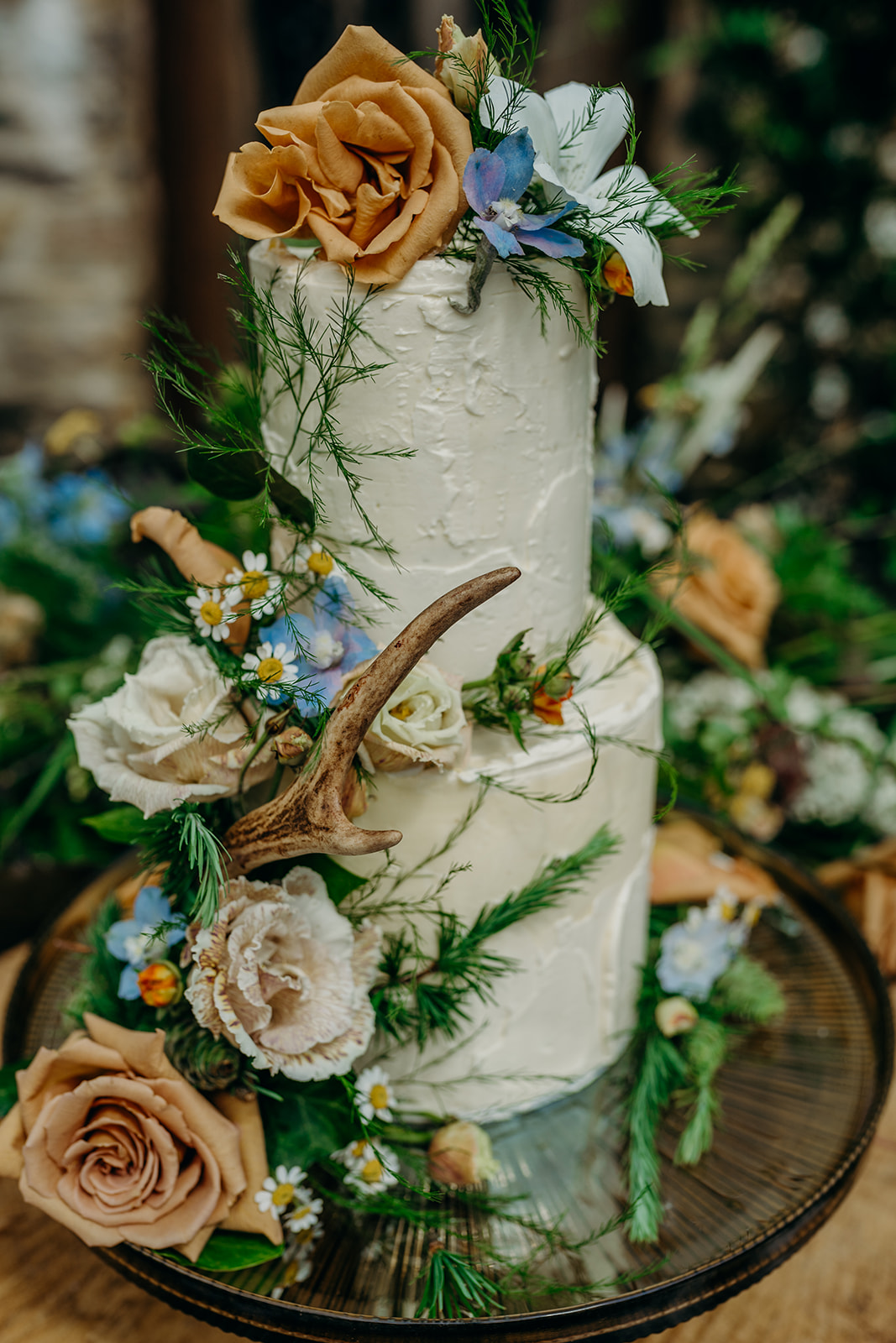 close up of two tier elopement wedding cake with rough buttercream icing on a dark lustre glass cake stand decorated with antlers and fresh flowers in caramel and pale blue colours