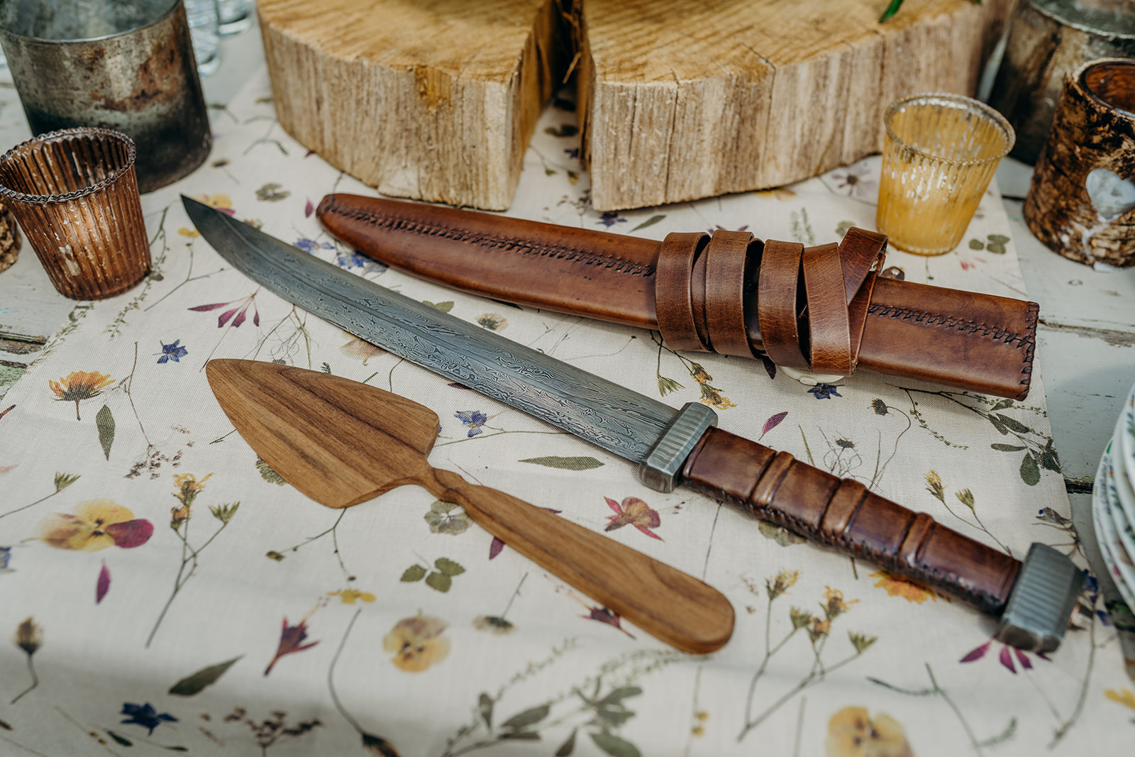 close up of hand made knife with leather sheath and handle by a wooden log slice on a pressed flower linen runner with rustic tealights
