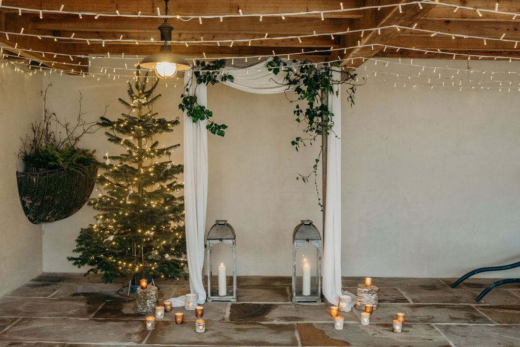 Wedding ceremony backdrop of draped arch with ivy next to a lit christmas tree with lanterns and candles at Ever After Devon