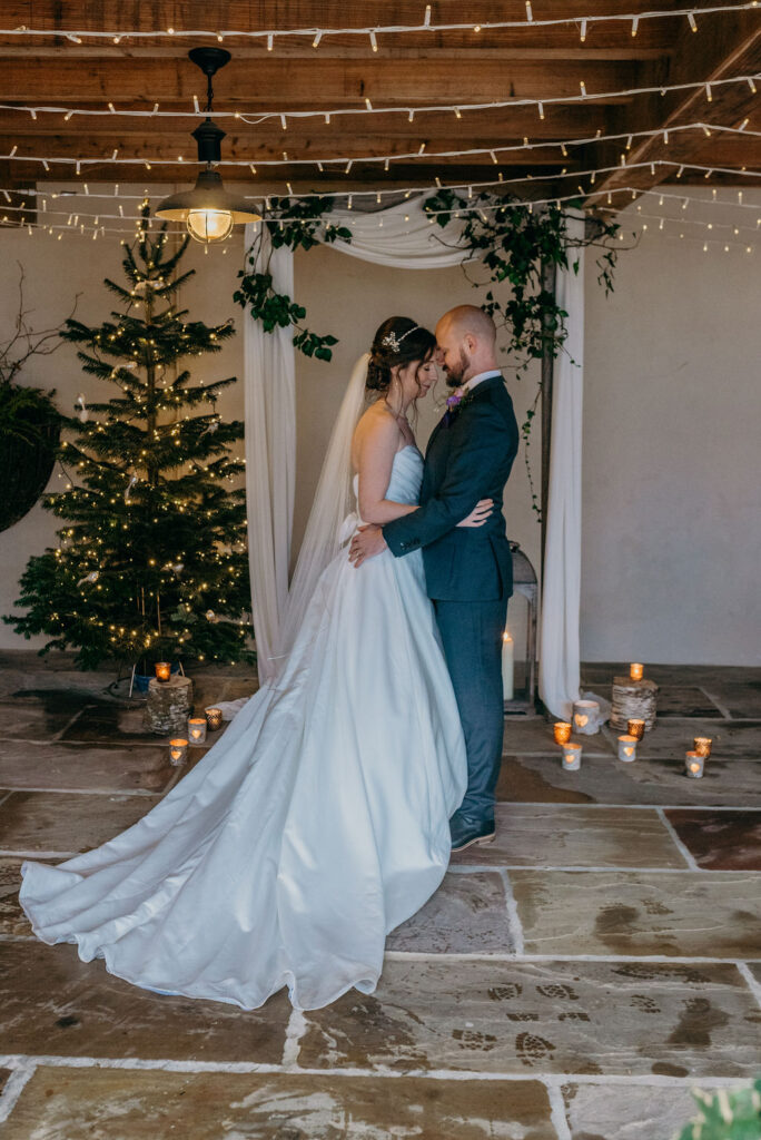 bride and groom in a wedding barn stood in front of a wooden arch draped with fabric and ivy next to a lit christmas tree