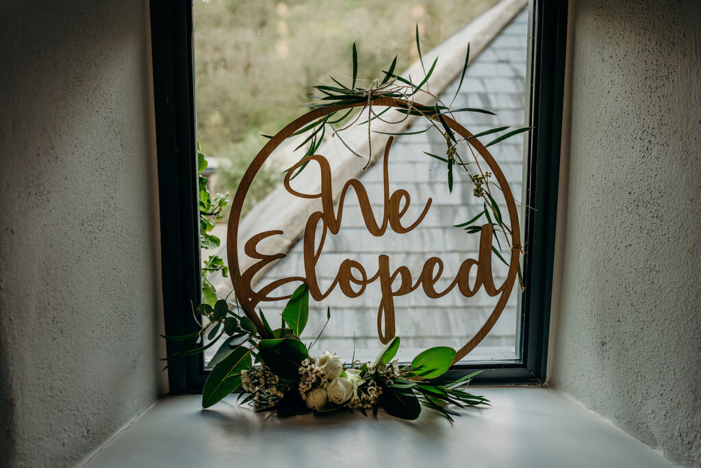wooden we eloped sign decorated with white flowers and green foliage leaning up against a window with slate roof in the background