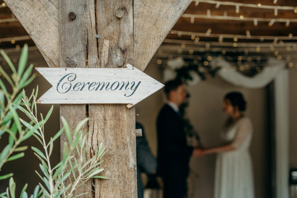 oak post with white hand painted Ceremony sign wedding couple unfocused in background