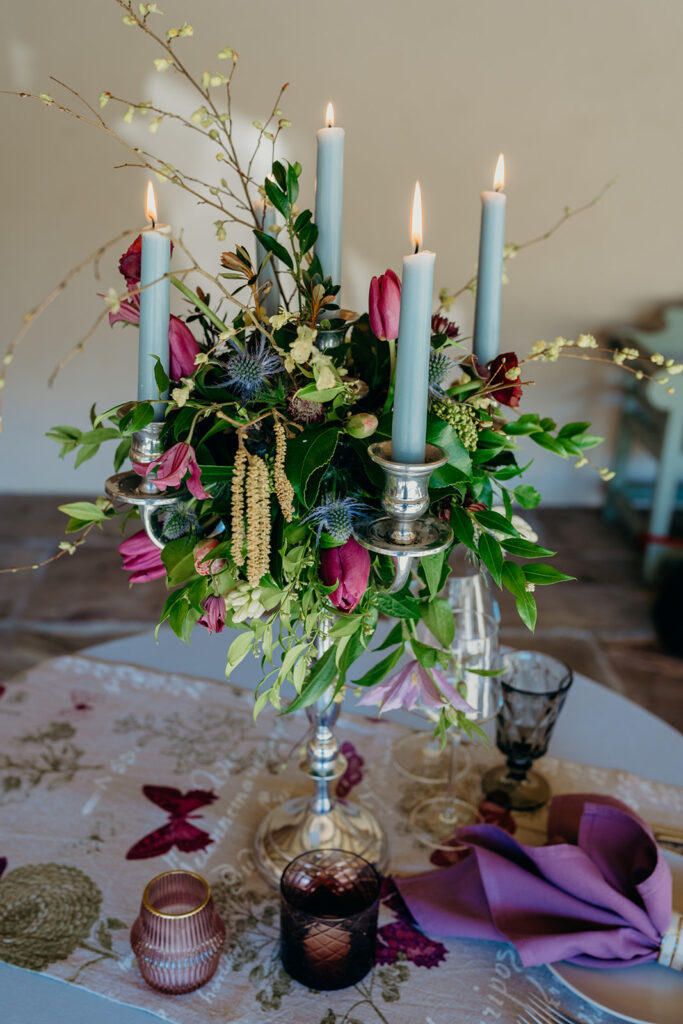 close up of silver candelabra decorated with flowers and grey candles on a table set with table runner and tealights