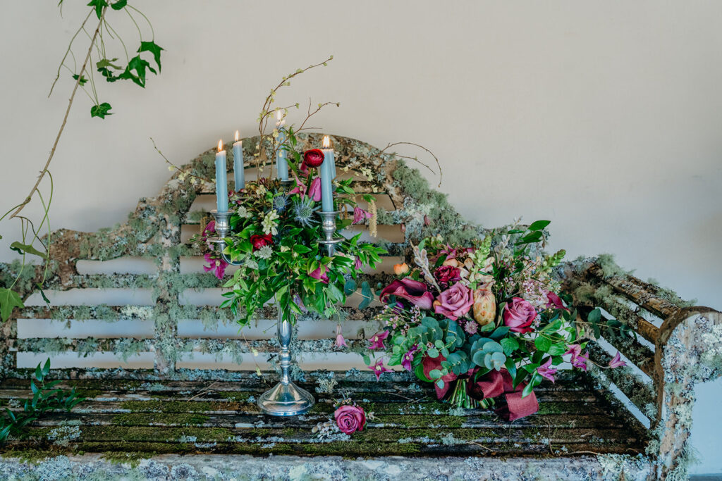 flower decorated silver candelabra with grey candles and a pink and purple bridal bouquet placed on a lichen covered Lutchens bench