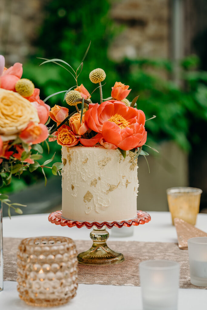 close up of mini wedding cake decorated with gold leaf and fresh coral peonies set on red and gold mini cake stand on a linen covered table with tea lights