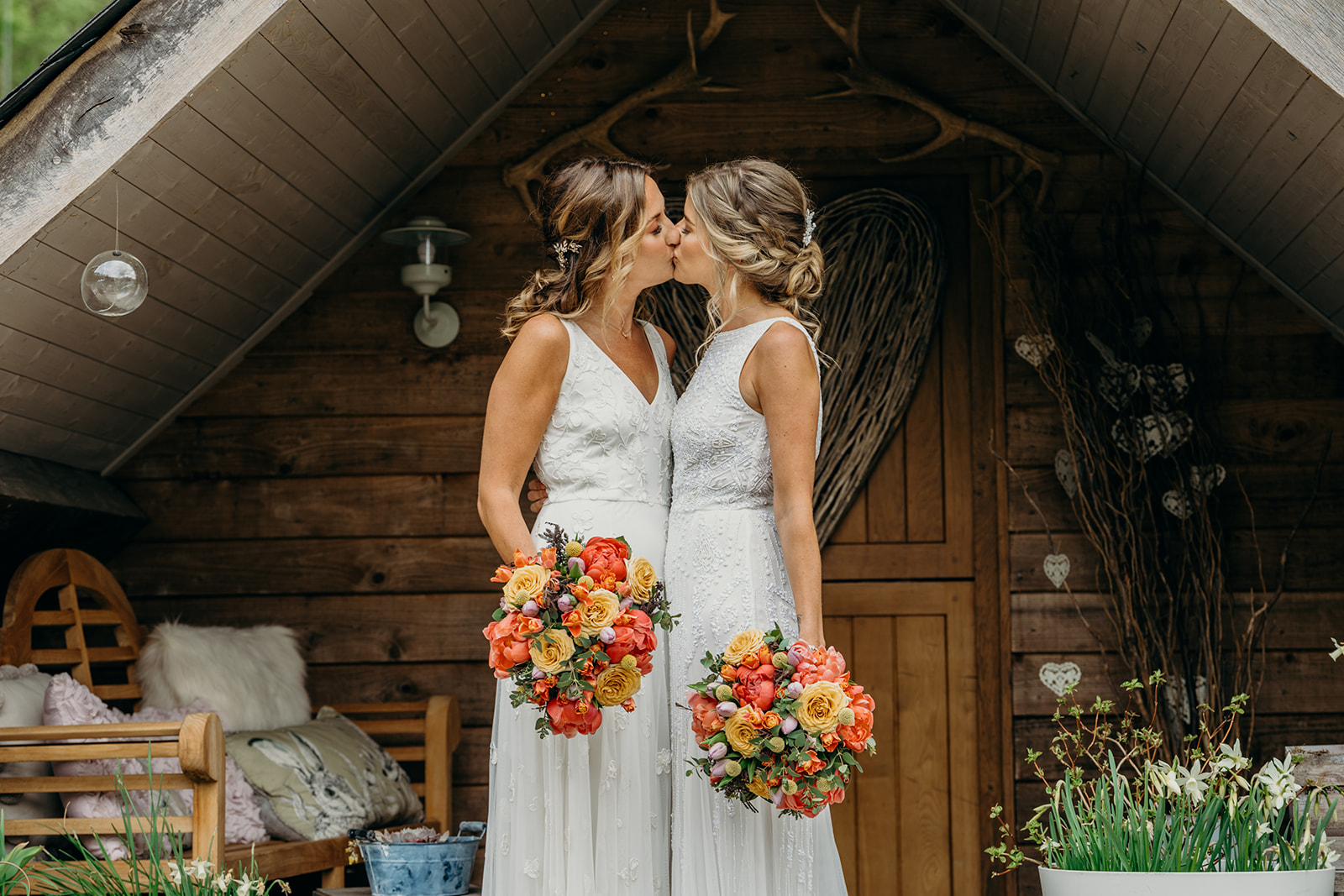 same sex elopement wedding with two blond white brides in mismatching white wedding dresses kissing in a rustic apex holding matching brightly coloured bridal bouquets in coral and yellow
