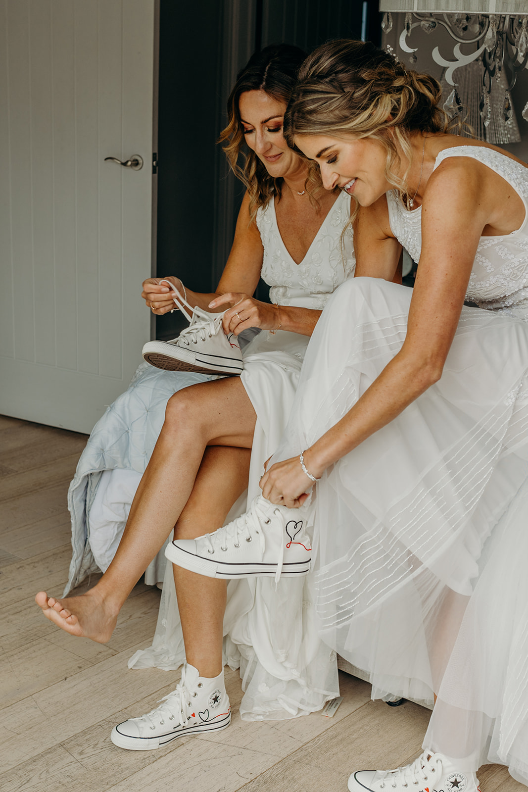 two white blonde brides in white wedding dresses sat on a bed putting on matching white converse trainers before getting married
