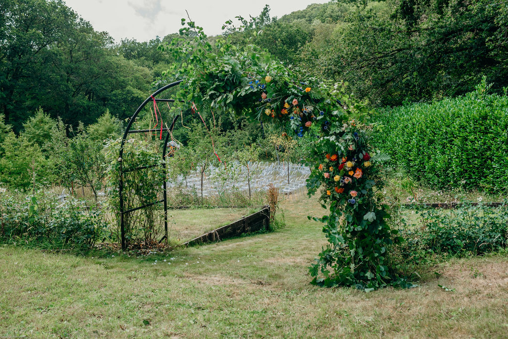 wedding ceremony set up in a Devon orchard with colourful flower arbour ribbons and ghost chairs