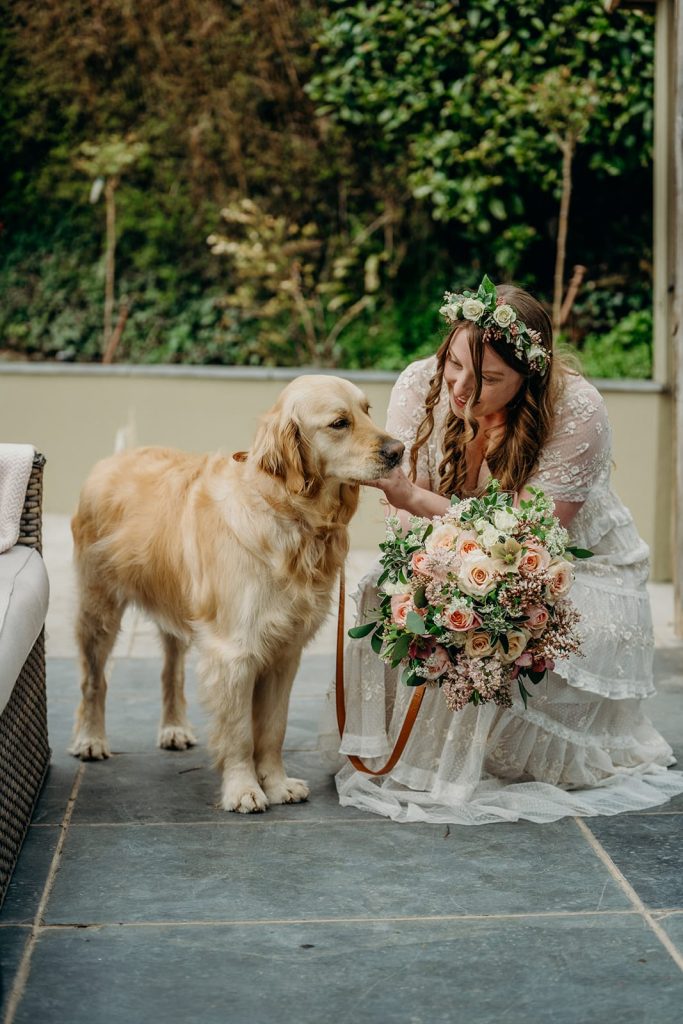 bride with bouquet kneeling down outside to pet a golden retriever dog on a slate patio