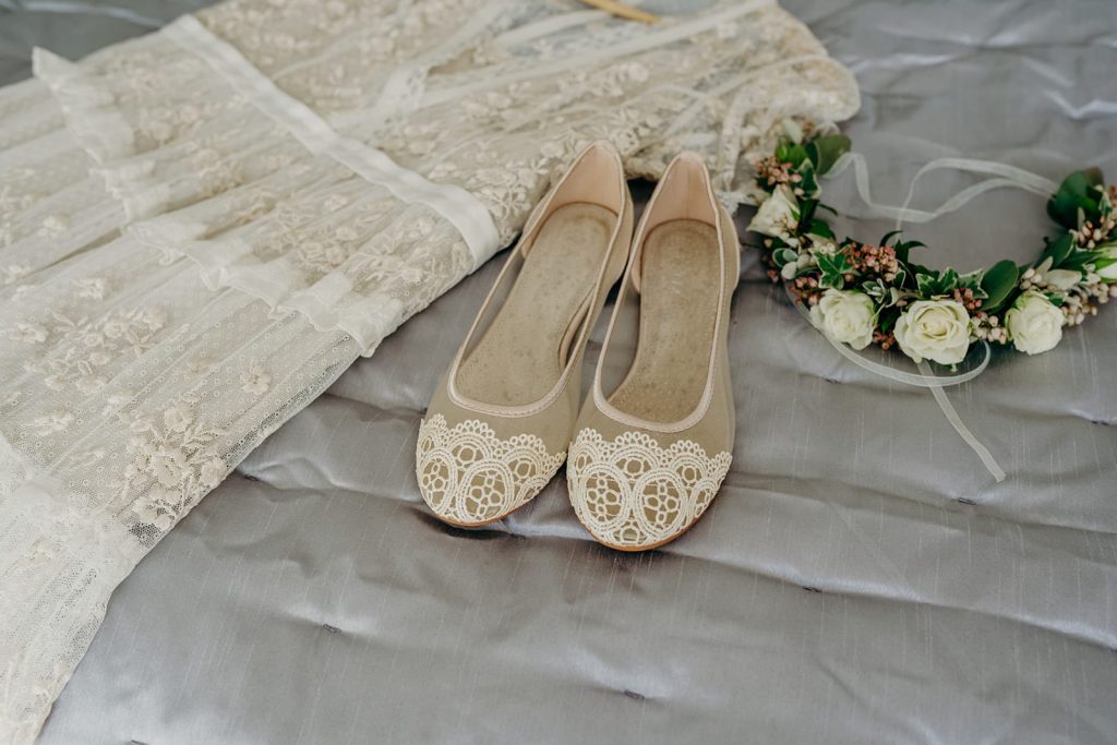 flat lay of lace wedding dress, ivory net & lace flat shoes and half head flower crown of ivory roses on a grey satin eiderdown