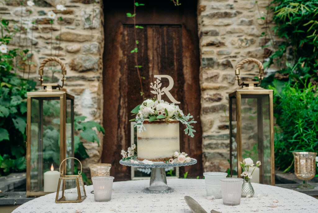mini semi naked wedding cake on a glass cake stand decorated with white fresh flowers and a wooden cake topper in front of a stone rustic wall with brass lanterns