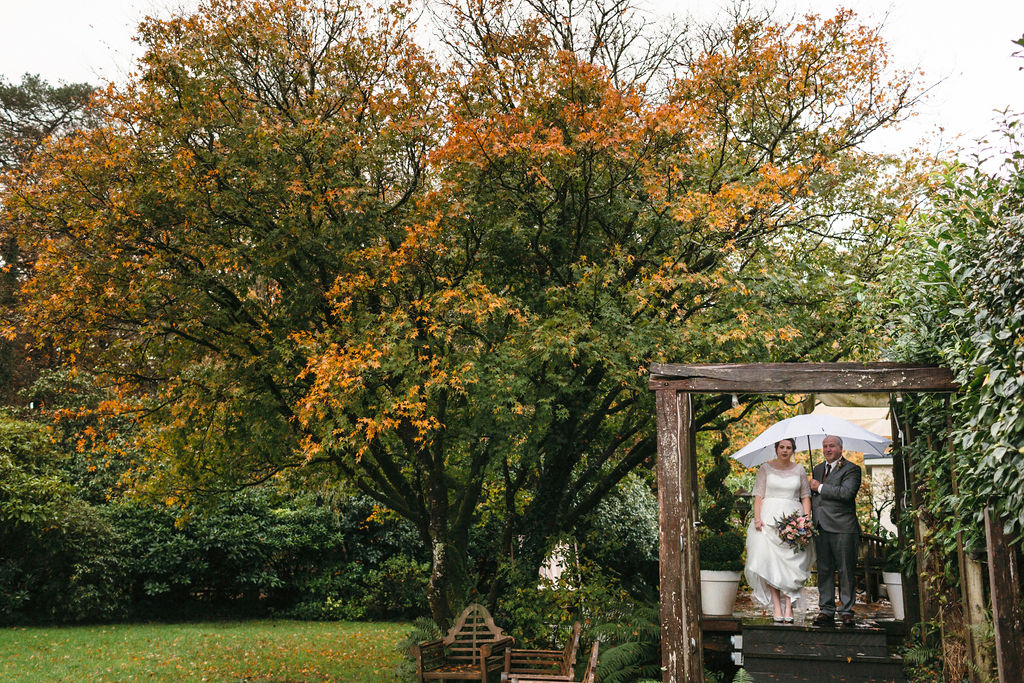 Bride and father stood on steps under a wide white umbrella with autumn colour trees waiting to walk into winter elopement wedding ceremony