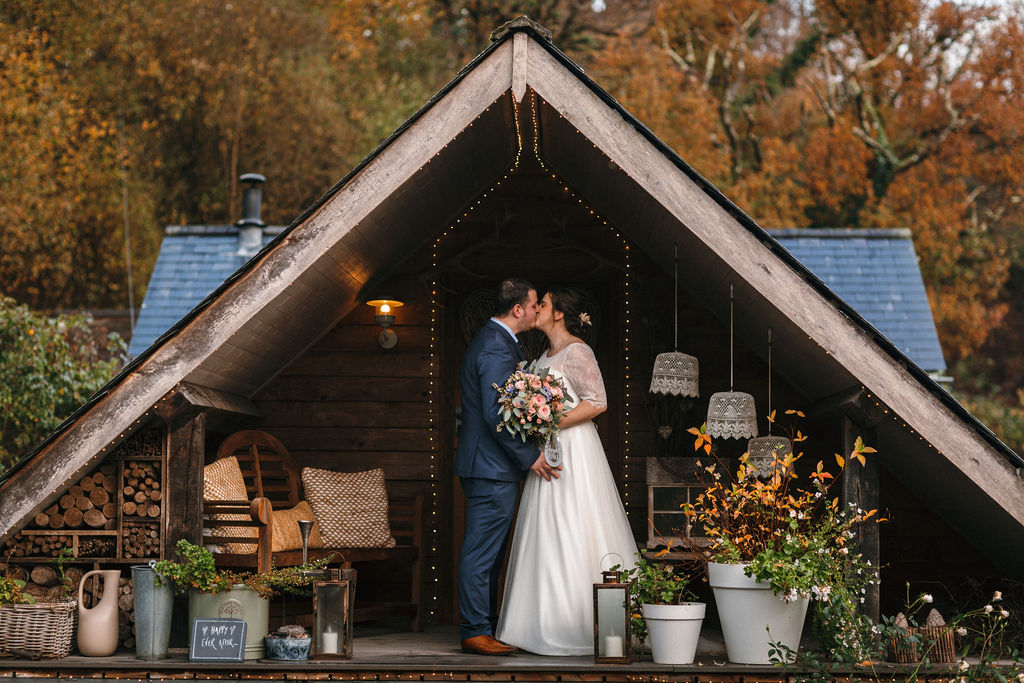 wedding couple stood kissing under wooden apex with autumn trees in background 