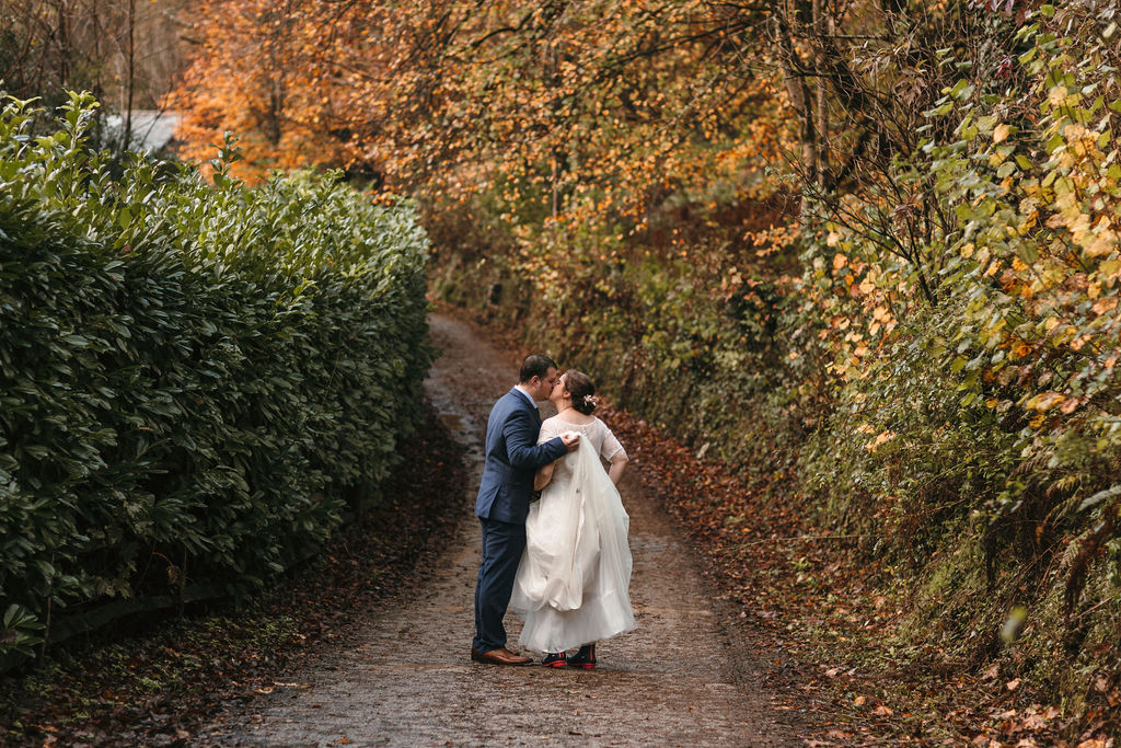 bride and groom kissing on country lane surrounded by trees in autumn leaf
