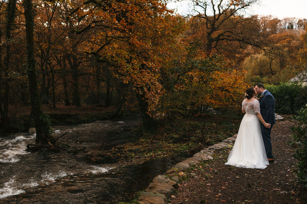 bride and groom stood by the banks of a river with backdrop of trees in autumn colours 