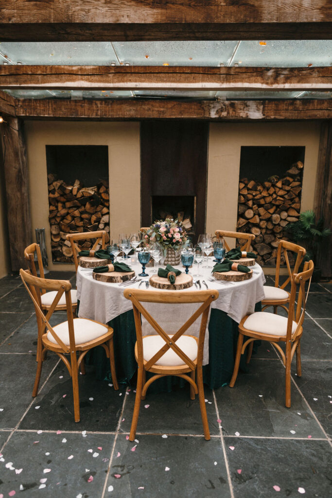 round wedding table dressed in dark green and natural linen cloths in front of log stores and fire place under oak framed glass roof, dark blue goblets and pink and blue bridal bouquet on the table