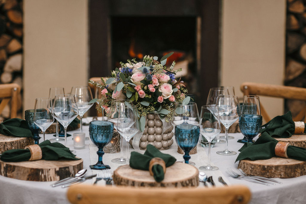 close up of wedding tablescape with log slice chargers, dark green linen napkins, pink and blue bouquet, dark blue wine goblets set in front of open fire