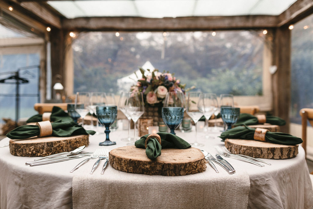 view of round wedding breakfast table dressed in natural linens, log slice chargers looking out through clear screen to gardens 