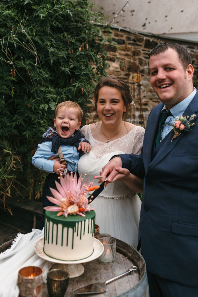 bride holding baby boy and groom stood in front of stone wall cutting mini wedding cake set on oak barrell decorated with dark green drip and pink dried flowers