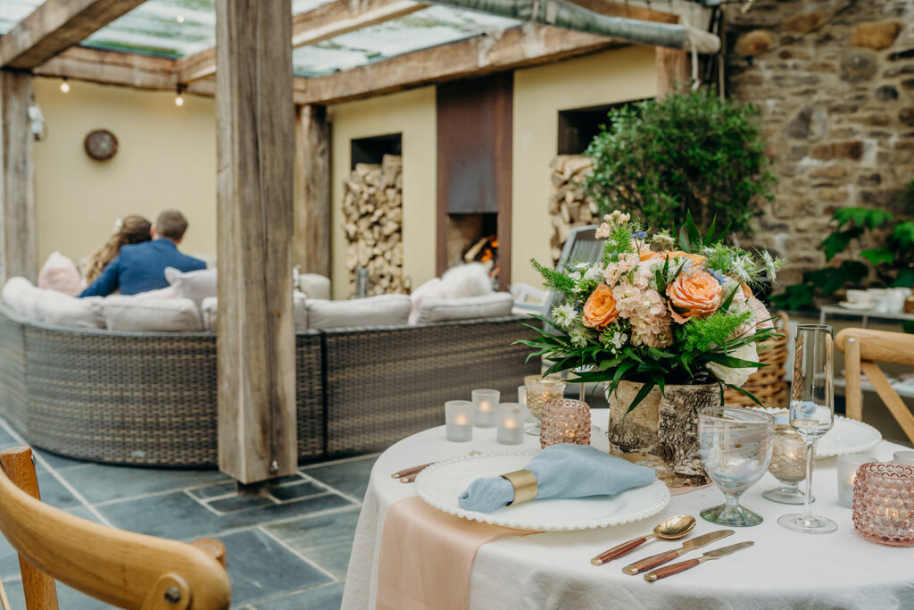 wedding table for 2 dressed in pink apricot and blue colours set up on a slate floor terrace with oak beams with back view of bride and groom seated on a sofa