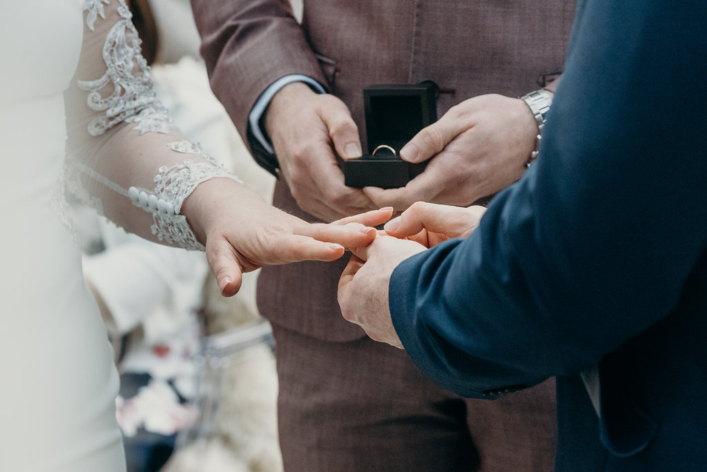 close up of bride and grooms hands with best man in background holding wedding ring box 