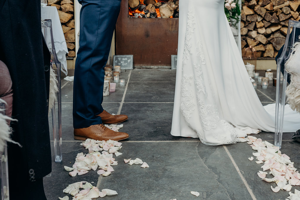 lower view of bride and groom stood facing each other in front of a log fire with pale pink rose petal aisle and guests sat on either side