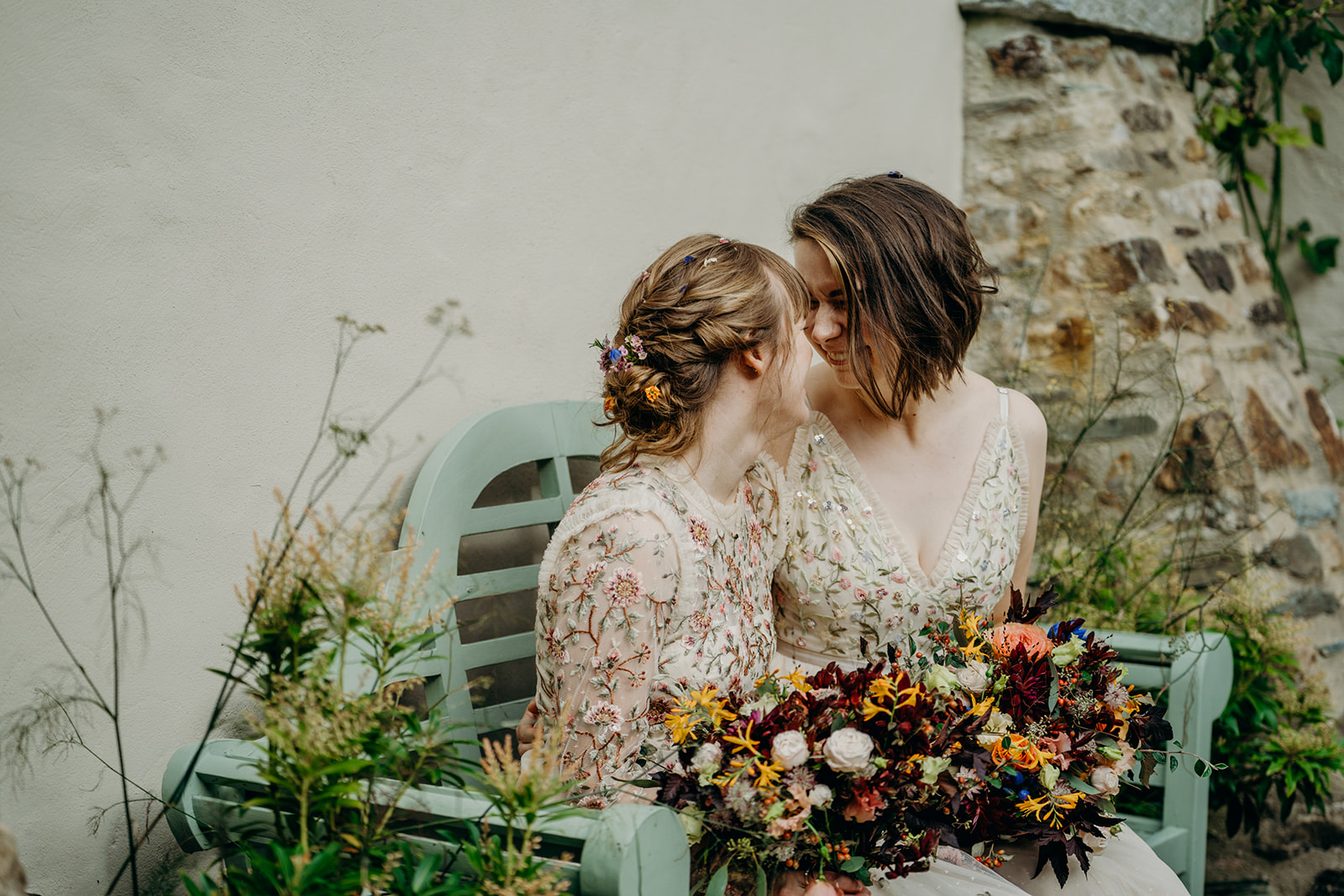 two white brides in mismatching embroidered wedding dresses sat on a green painted lutyens bench looking at each other and smiling with matching bouquets in dark colours and orange on their laps