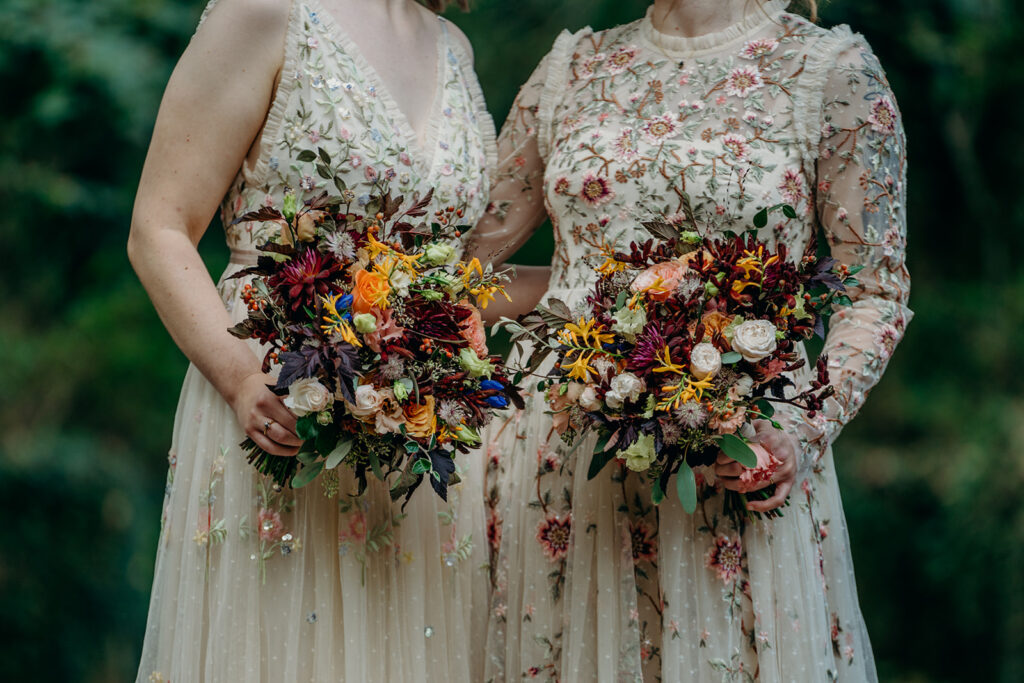 mid body shot of two brides wearing flower embroidered wedding dress carrying bouquets of orange and dark red
