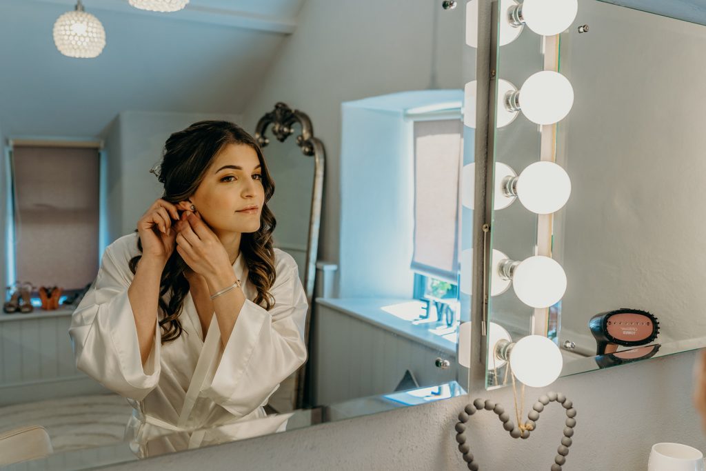 bride with long dark hair and a white satin robe putting on earrings in front of a mirror